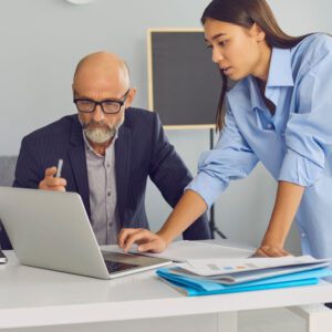 Technische Projektbesprechung zwischen älterem Mann im Anzug und junger Frau am Laptop in modernem Büro.