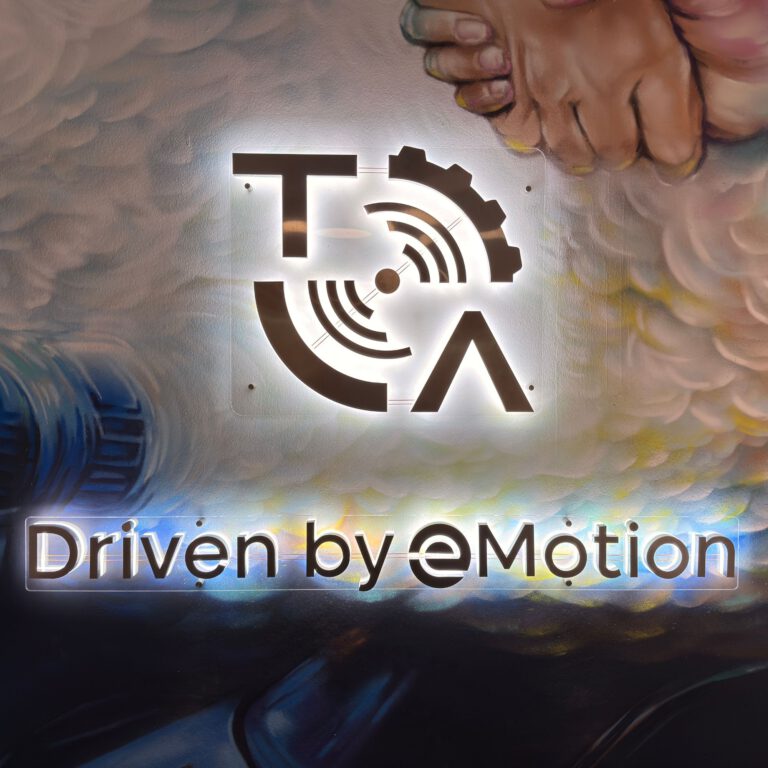 Leuchtendes Logo von Technische Antriebselemente mit dem Schriftzug „Driven by eMotion“ vor einer künstlerisch gestalteten Wand, die technische und menschliche Elemente wie Zahnräder und zwei sich haltende Hände vereint.