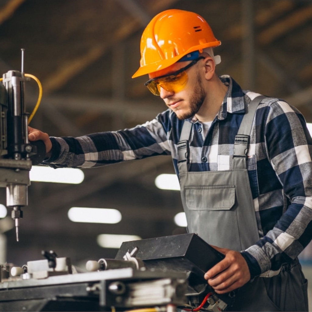 Ein junger Industriearbeiter mit orangefarbenem Schutzhelm und gelber Schutzbrille bedient konzentriert eine Maschine in einer Fabrikhalle.