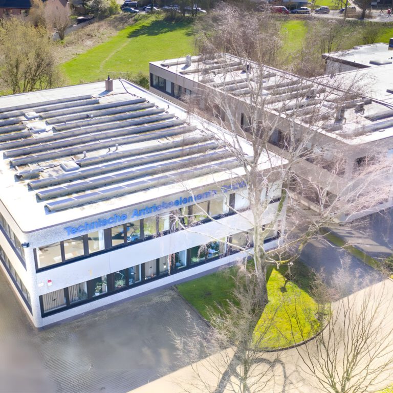 Luftaufnahme des Firmengebäudes von Technische Antriebselemente GmbH in Hamburg. Das moderne zweistöckige Büro- und Lagergebäude mit großflächigen Fenstern und installierter Photovoltaikanlage auf dem Dach liegt in einem grünen Gewerbegebiet. Im Vordergrund sind Einfahrt und Bäume zu sehen, im Hintergrund Grünflächen und weitere Gebäude.