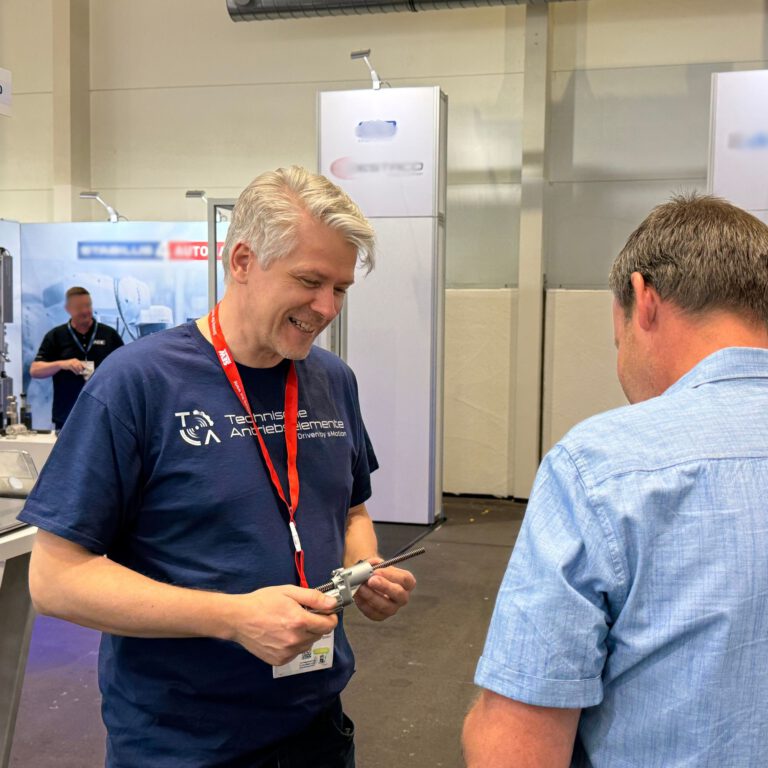 Ein Mitarbeiter von Technische Antriebselemente erklärt auf einer Messe einem Besucher ein mechanisches Bauteil, ein Spindelhubgetriebe. Beide stehen an einem Messestand, im Hintergrund sind weitere Aussteller zu sehen.